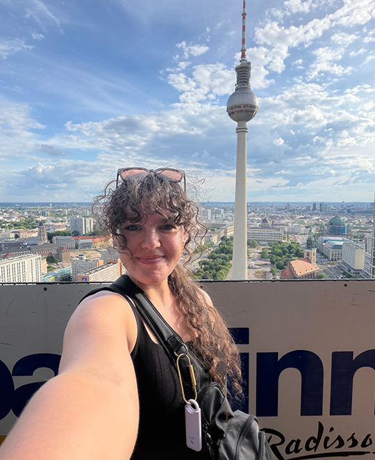Elizabeth Gruen taking a selfie outdoors with the Berlin TV Tower (Berliner Fernsehturm) visible in the background.