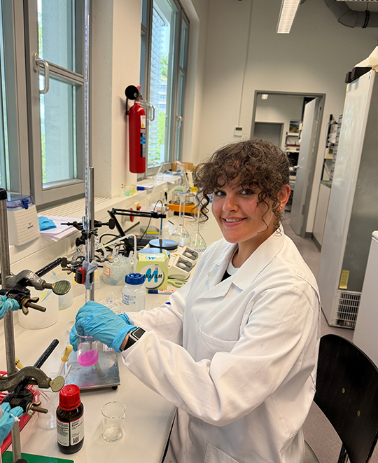Elizabeth Gruen wearing safety glasses and holding lab glassware while examining a liquid sample in a laboratory.