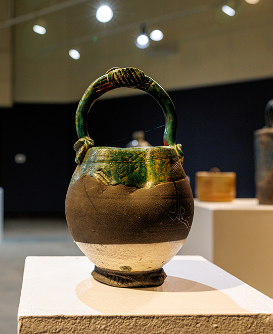 Ceramic teapot with a dark woven handle, glossy glazed lid, and an earthy stoneware body displayed on a white pedestal.