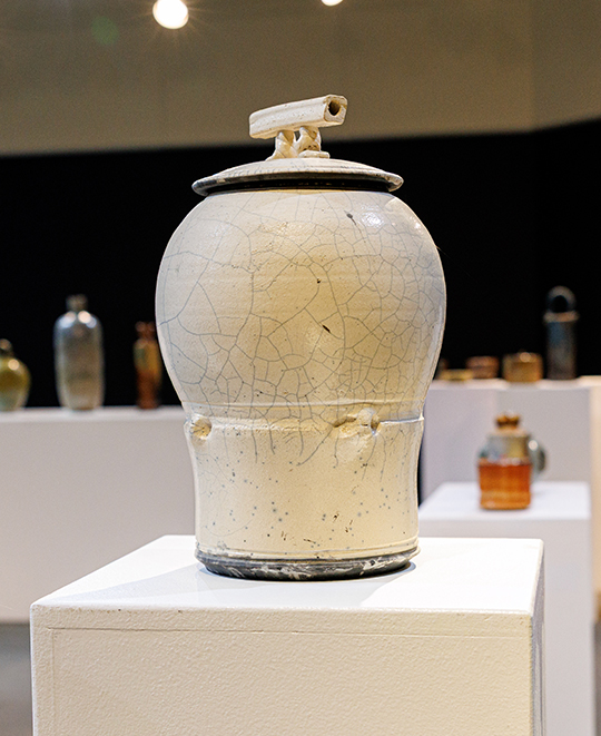 Tall, rounded ceramic jar with an off-white glazed body, dark neck, and broad base displayed on a white pedestal in an art gallery.