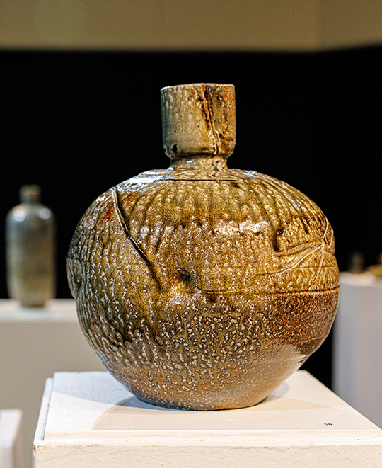 Rounded, earthy ceramic jar with a dark spout and speckled brown glaze displayed on a white pedestal in an art gallery.