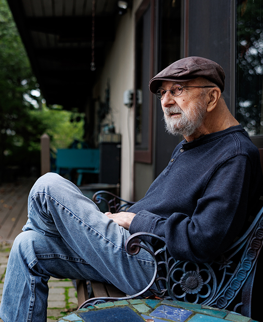 Bruce Chesser sitting outdoors on a wooden bench, wearing a cap and jacket while looking off to the side.