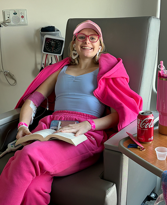 Cassandra Kurek smiling while sitting in a chair during medical treatment, wearing a bright pink outfit.