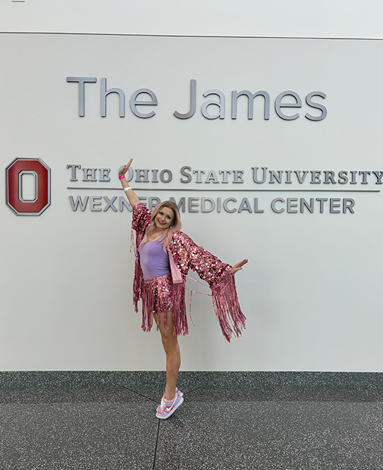 Cassandra Kurek smiling in a bright pink outfit while standing in front of a wall featuring "The James" sign at The Ohio State University Comprehensive Cancer Center.