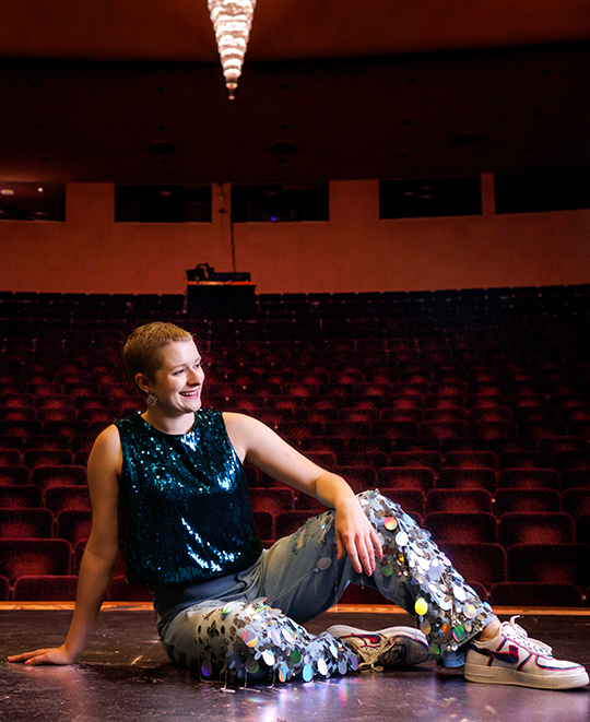 Cassandra Kurek wearing a blue sequined outfit, sitting on the floor of a stage inside an auditorium.