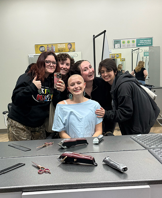 Cassandra Kurek seated in a chair looking toward the mirror as friends lean in closely on either side, all smiling.