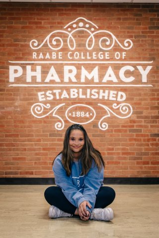 Photo of Veronica seated on the floor with pharmacy sign above
