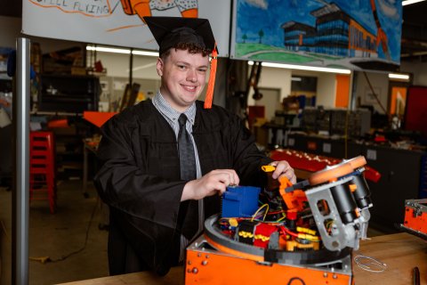 Photo of Kaden working on a robot while wearing his graduation cap and gown