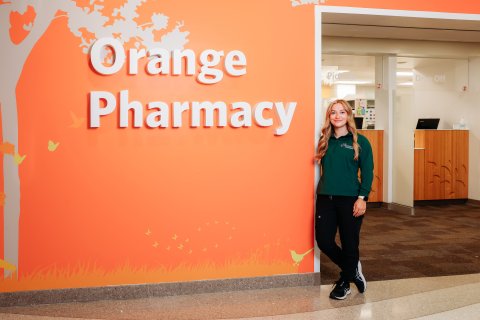Photo of Ashely standing next to orange wall with text that says orange pharmacy on it