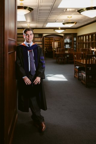 Andrea wearing his graduation gown leaning on the door to the law library