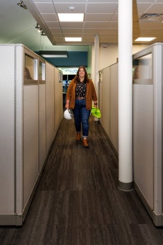Mollie in a office walking down a cubicle hallway holding a hard hat and a reflective vest
