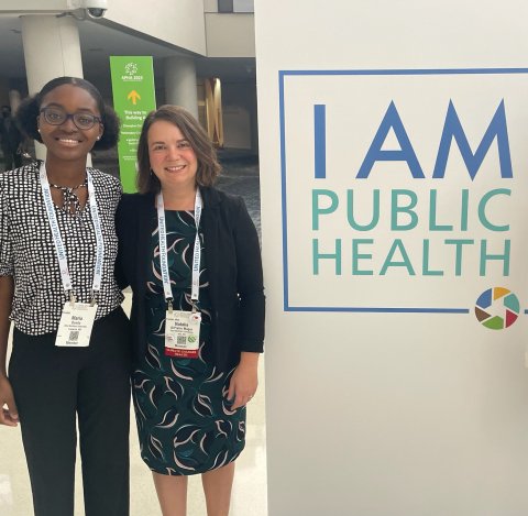 Photo of Maria and her professor next to a sign that says I am public health