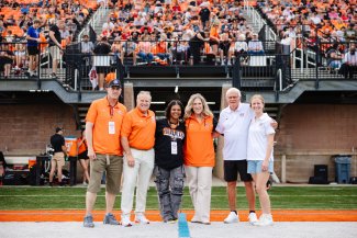 Six people stand on a football field with a crowd in the stands behind them