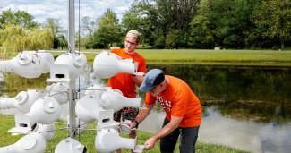 Two people wearing orange shirts check a cluster of white gourd birdhouses mounted on a metal pole next to a pond.